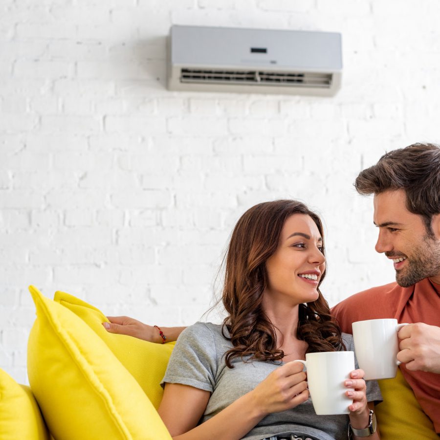 happy couple with cups sitting on sofa under air conditioner at home
